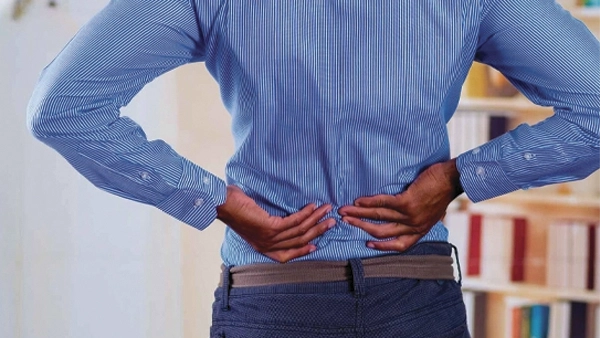 Person standing indoors with both hands placed on the lower back, highlighting the lumbar area against a blurred background.